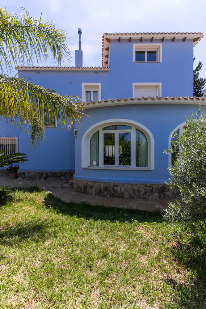 A two-story blue house with a stone base, arched windows, and a tiled roof, set behind a green lawn and palm tree.
