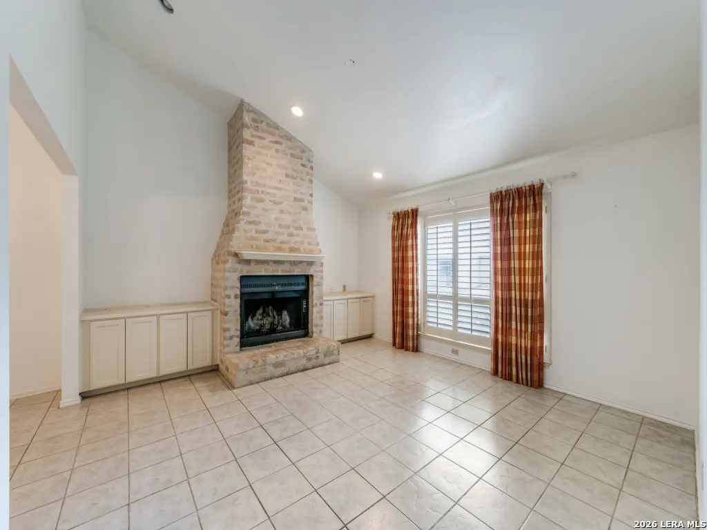 A living room with a brick fireplace, white walls, and tile flooring. Red plaid curtains frame a window with white shutters.