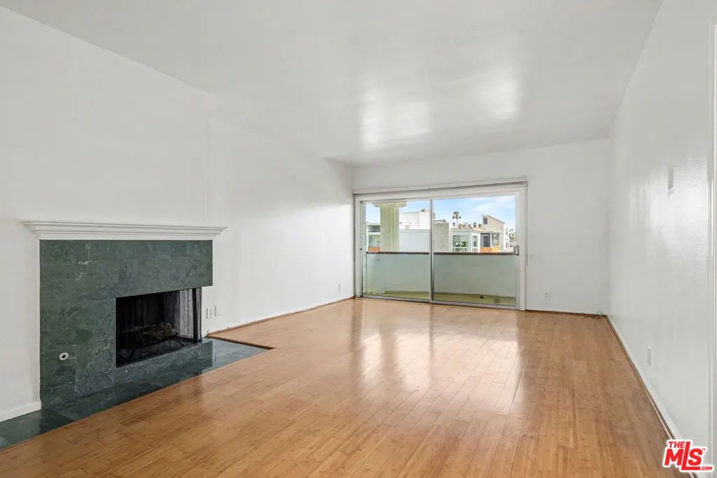 Bright, empty living room with hardwood floors, white walls, and a green marble fireplace. Sliding glass doors lead to a balcony.