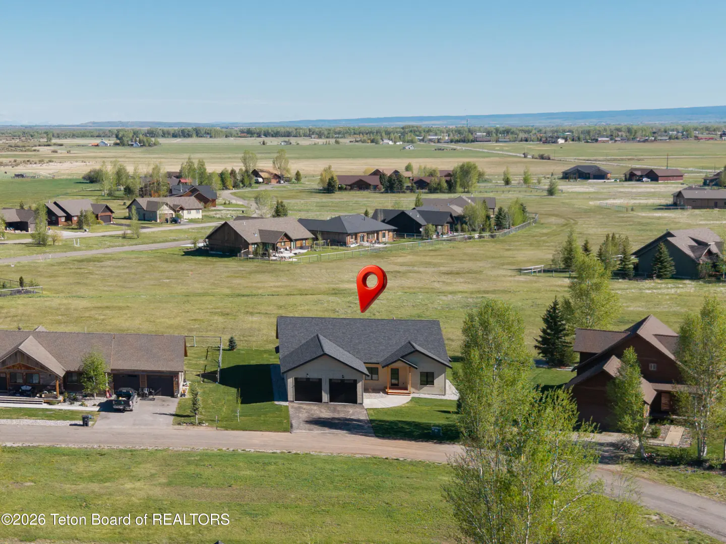 Aerial view of a gray house with a red location pin above it, in a green field with other houses.