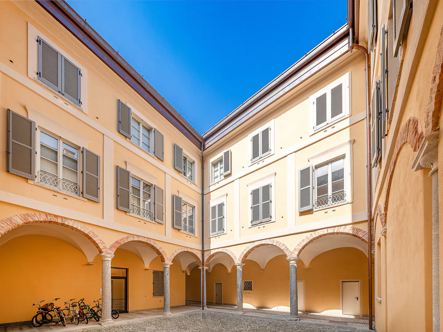 Courtyard view of a yellow building with arched walkways, gray shutters, and a cobblestone ground. Bicycles are parked on the left.