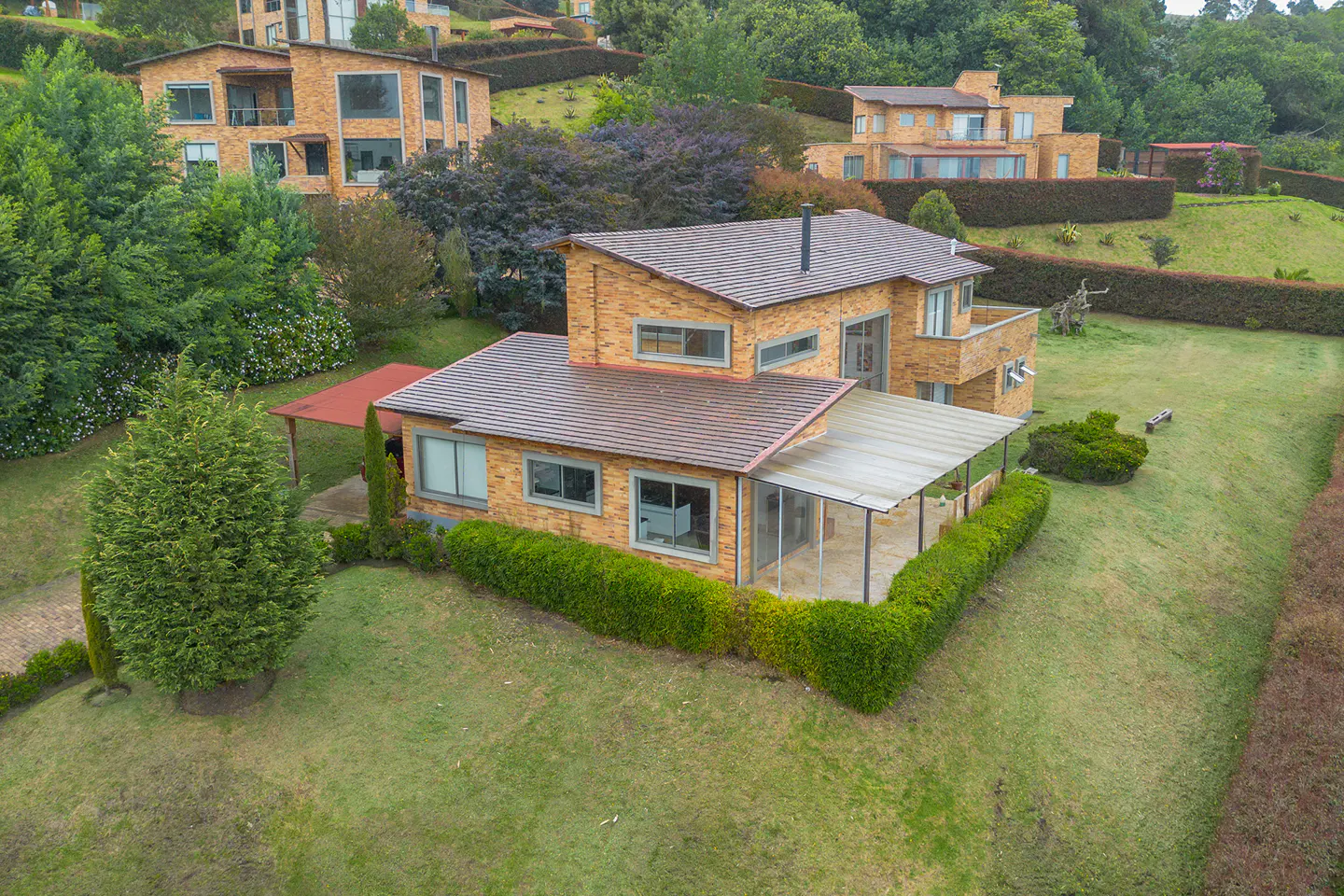 Aerial view of a two-story brick house with a brown roof, surrounded by green grass and hedges on a sloped lawn.