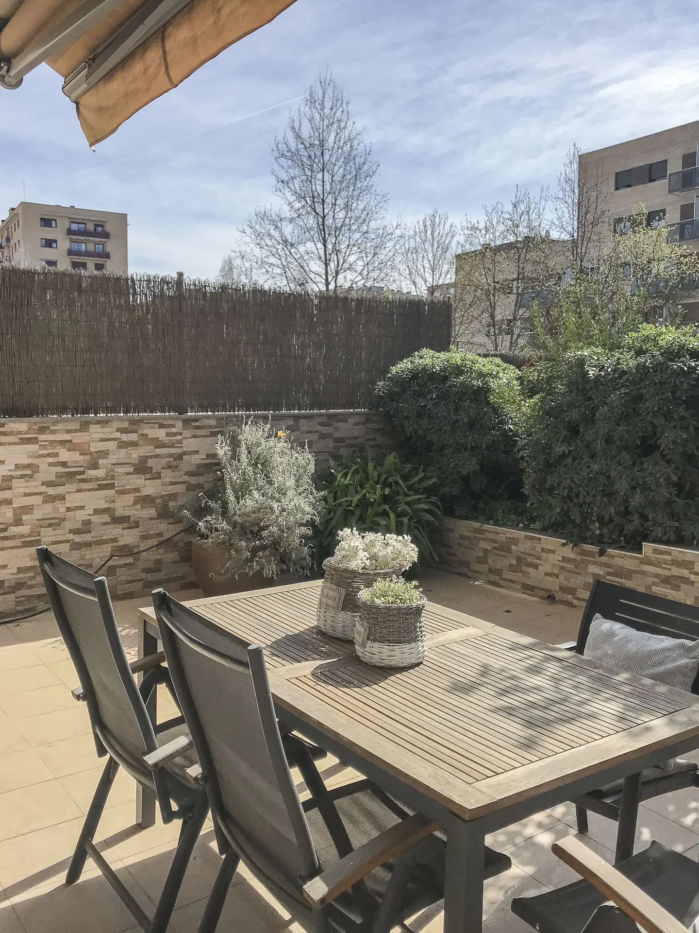 Outdoor patio with a wooden table, chairs, and potted plants. A woven fence and greenery provide privacy. Buildings are visible in the background.