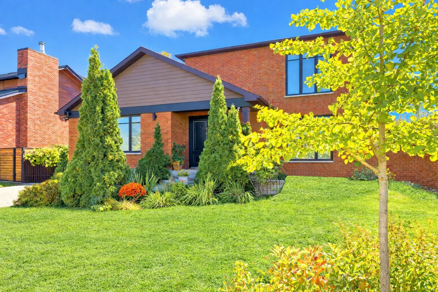 Modern red brick house with a brown gabled roof, lush green lawn, manicured evergreen trees, and bright blue sky.