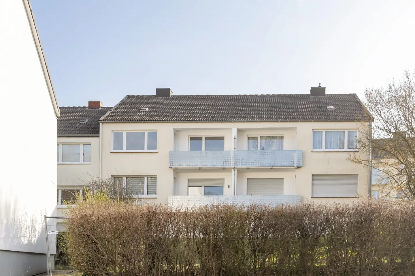 A three-story apartment building with balconies, a gray roof, and a hedge in front.
