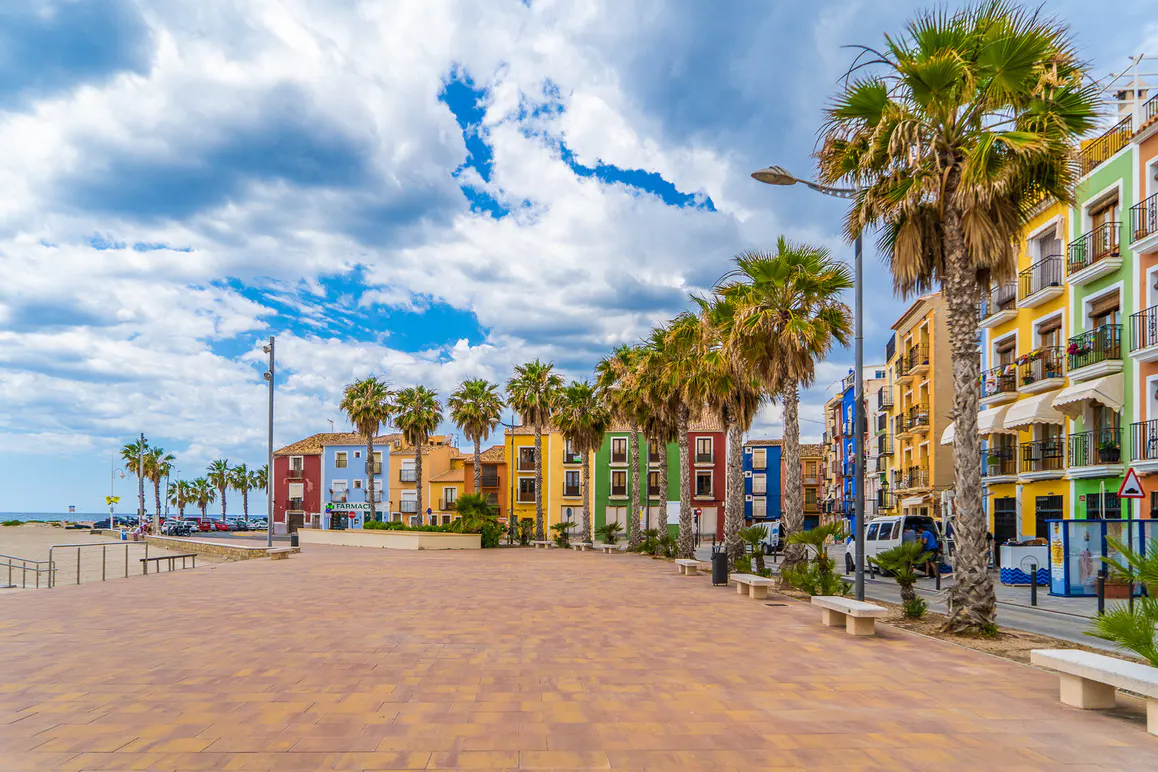 Colorful buildings line a plaza with palm trees under a cloudy sky. A beach is visible in the distance.