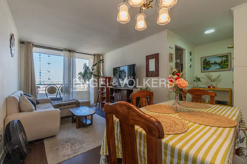 Living room with a dining table set with placemats and a striped tablecloth. A beige sofa sits near a window with city views. A TV and plants are in the background.