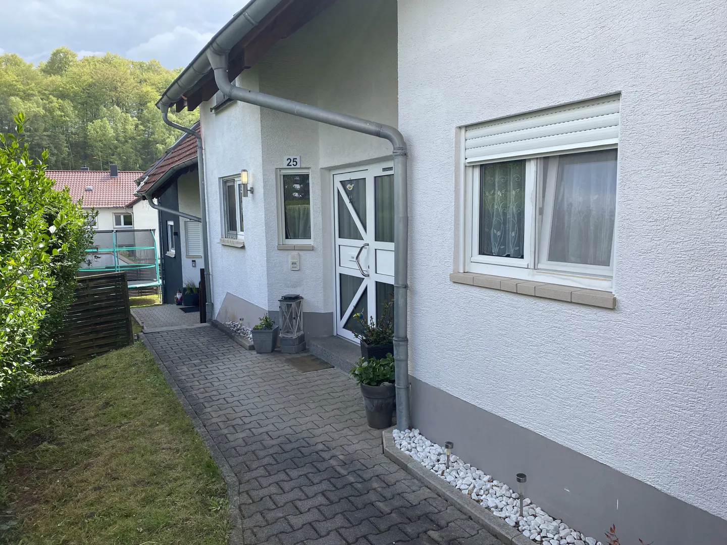 Exterior view of a white house with a gray brick walkway leading to the front door. A window with a white frame is on the right. The house number "25" is visible above the door.