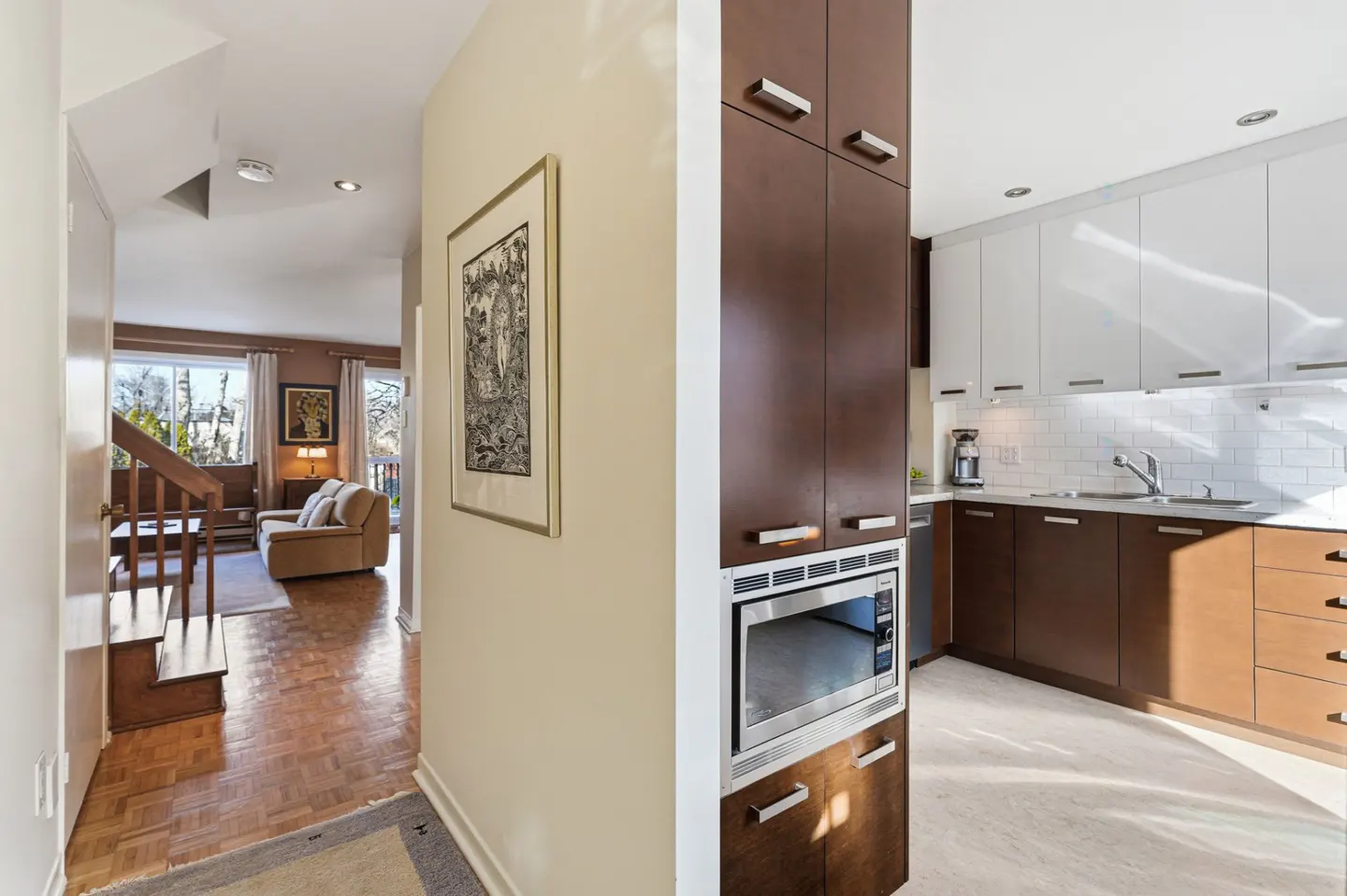 Hallway view into a living room with stairs and a kitchen with brown and white cabinets.