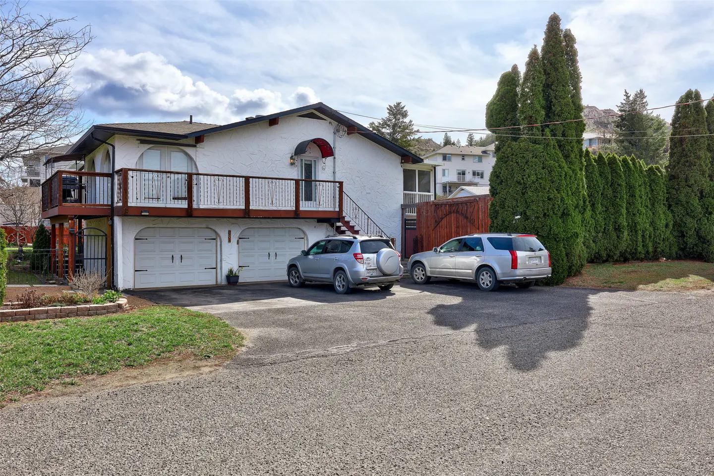 A two-story white house with a brown roof, two garage doors, a balcony, and two silver cars parked in front.