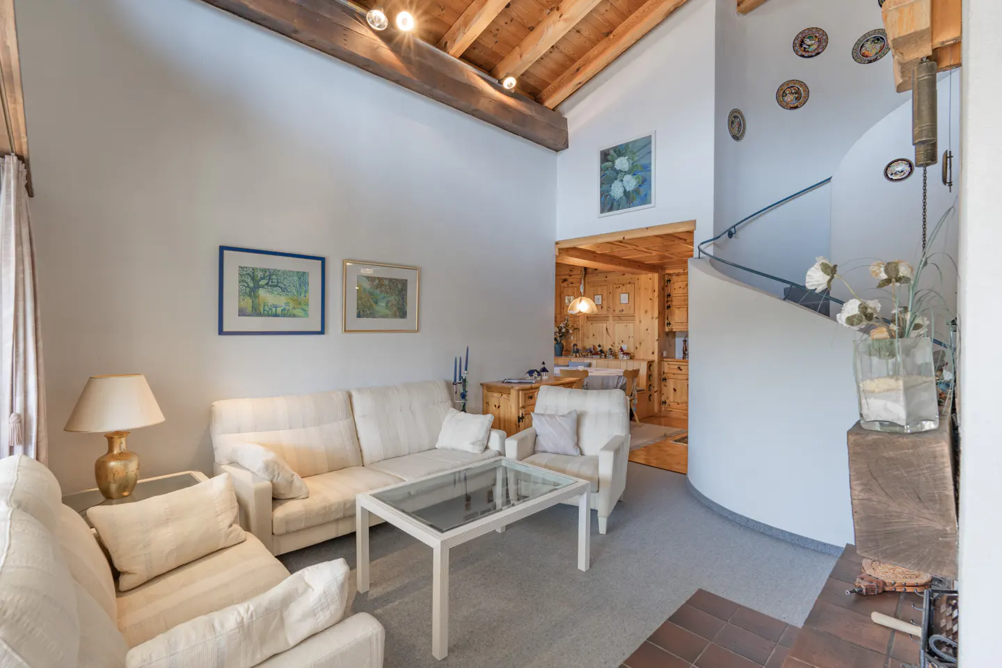 Living room with white sofas, glass table, and wooden ceiling beams. A curved staircase leads up, decorated with plates.