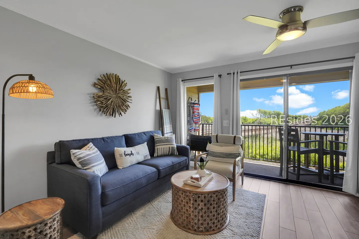 Living room with blue sofa, patterned pillows, and a round wooden coffee table. Sliding glass doors lead to a balcony with a view.