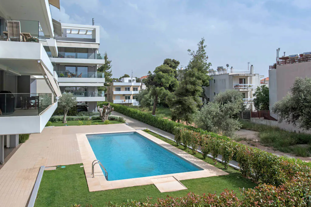 A rectangular blue swimming pool is surrounded by green grass and hedges, with modern white buildings in the background.
