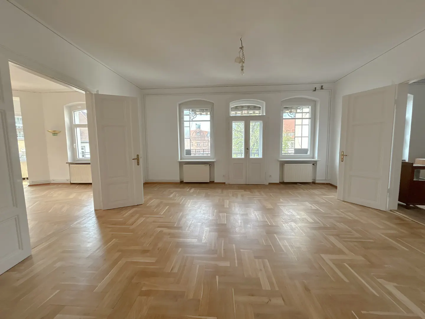 Empty room with herringbone wood floors, white walls, and three windows. Two open white doors lead to other rooms. Radiators under windows.