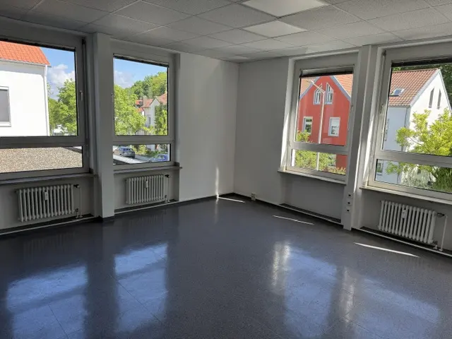 Empty room with gray floor, white walls, and four windows showing buildings and trees outside. Radiators are under each window.