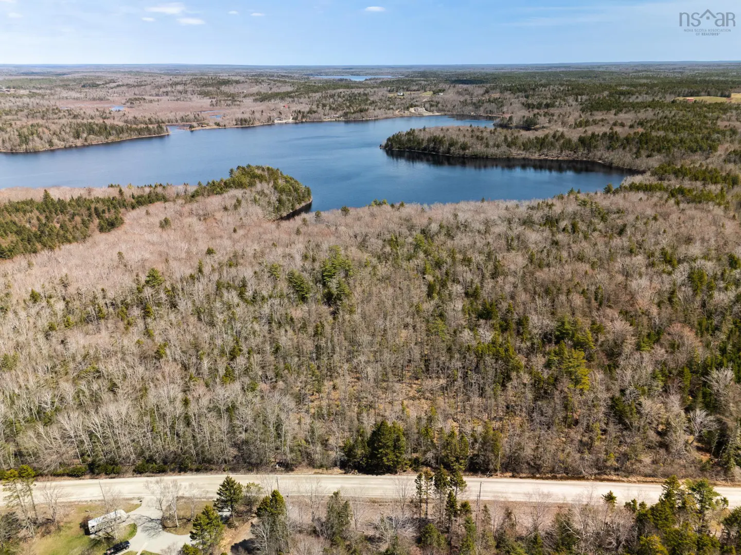 Aerial view of a lake surrounded by trees, with a road and a few houses visible in the foreground.