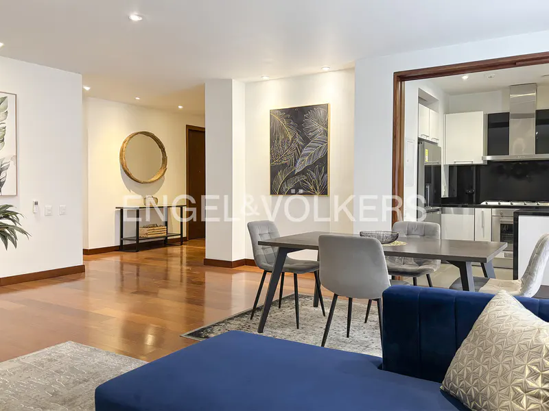 A modern living and dining area with hardwood floors, a blue sofa, and a dining table with gray chairs. A kitchen is visible in the background.