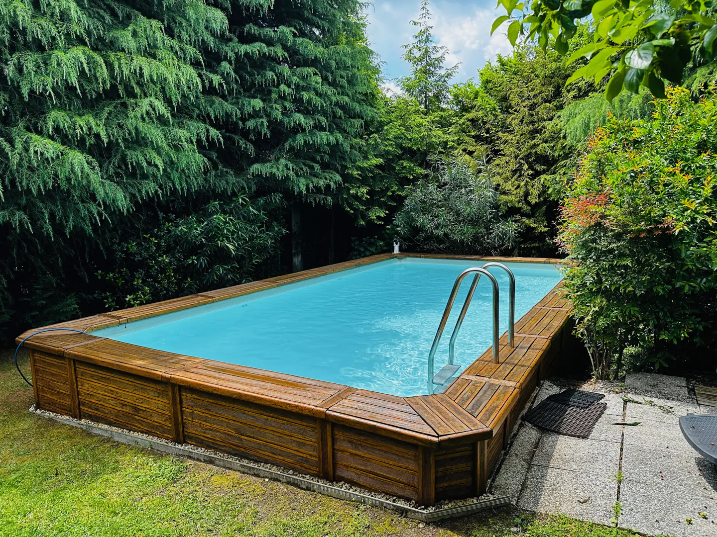 Backyard swimming pool with wooden deck and metal ladder, surrounded by lush green trees and grass.