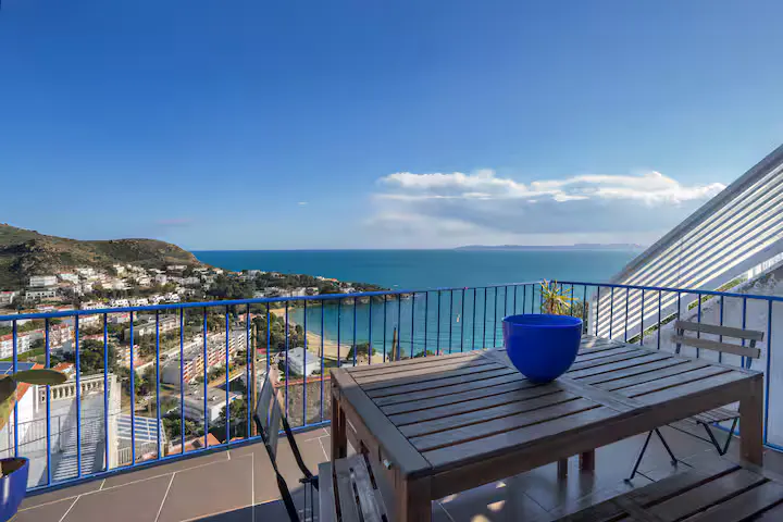 Wooden patio table with a blue bowl overlooking a coastal town, sandy beach, and the blue ocean under a clear sky.