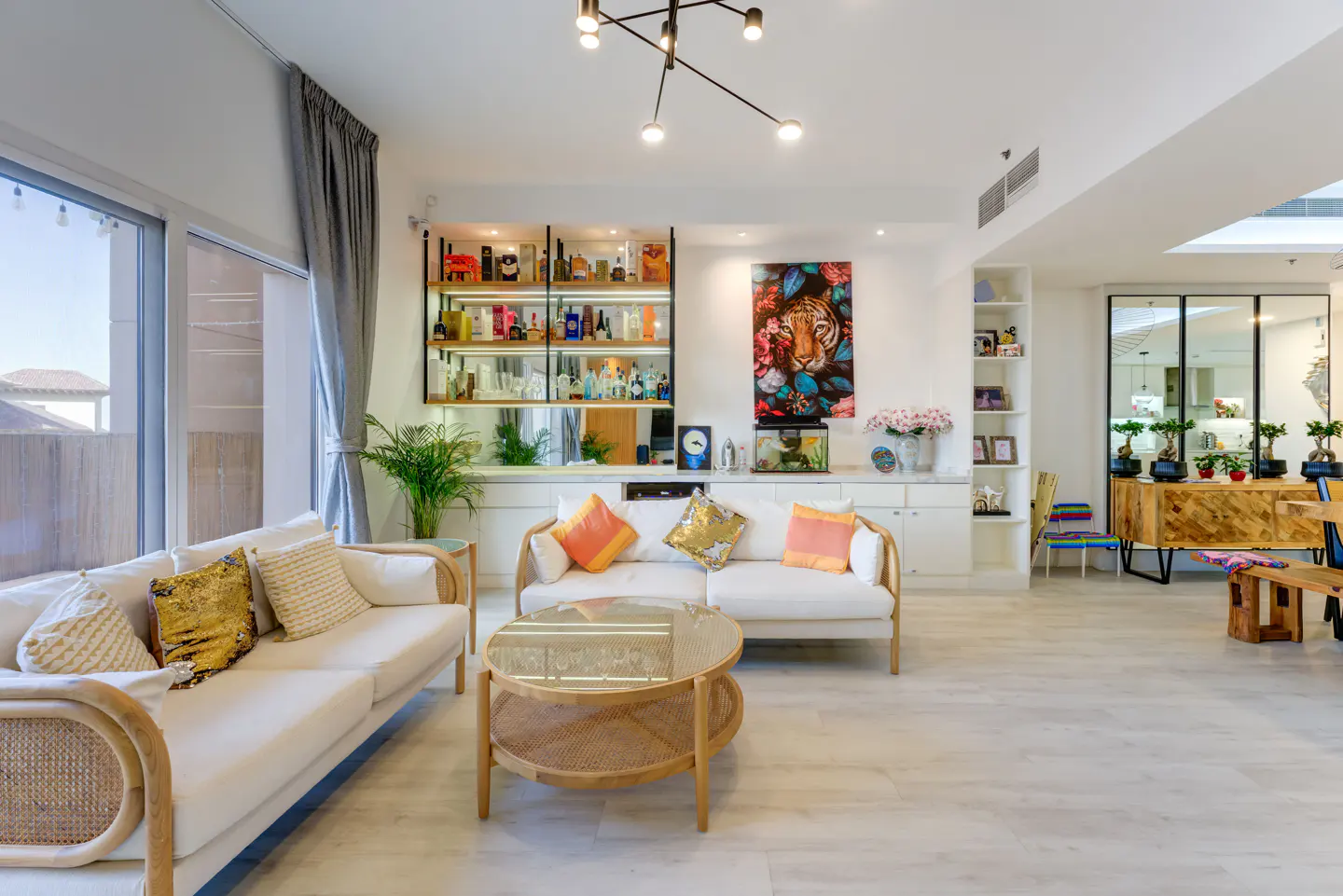 Bright living room with white sofas, rattan coffee table, and a bar with liquor bottles. A tiger painting hangs above a fish tank.