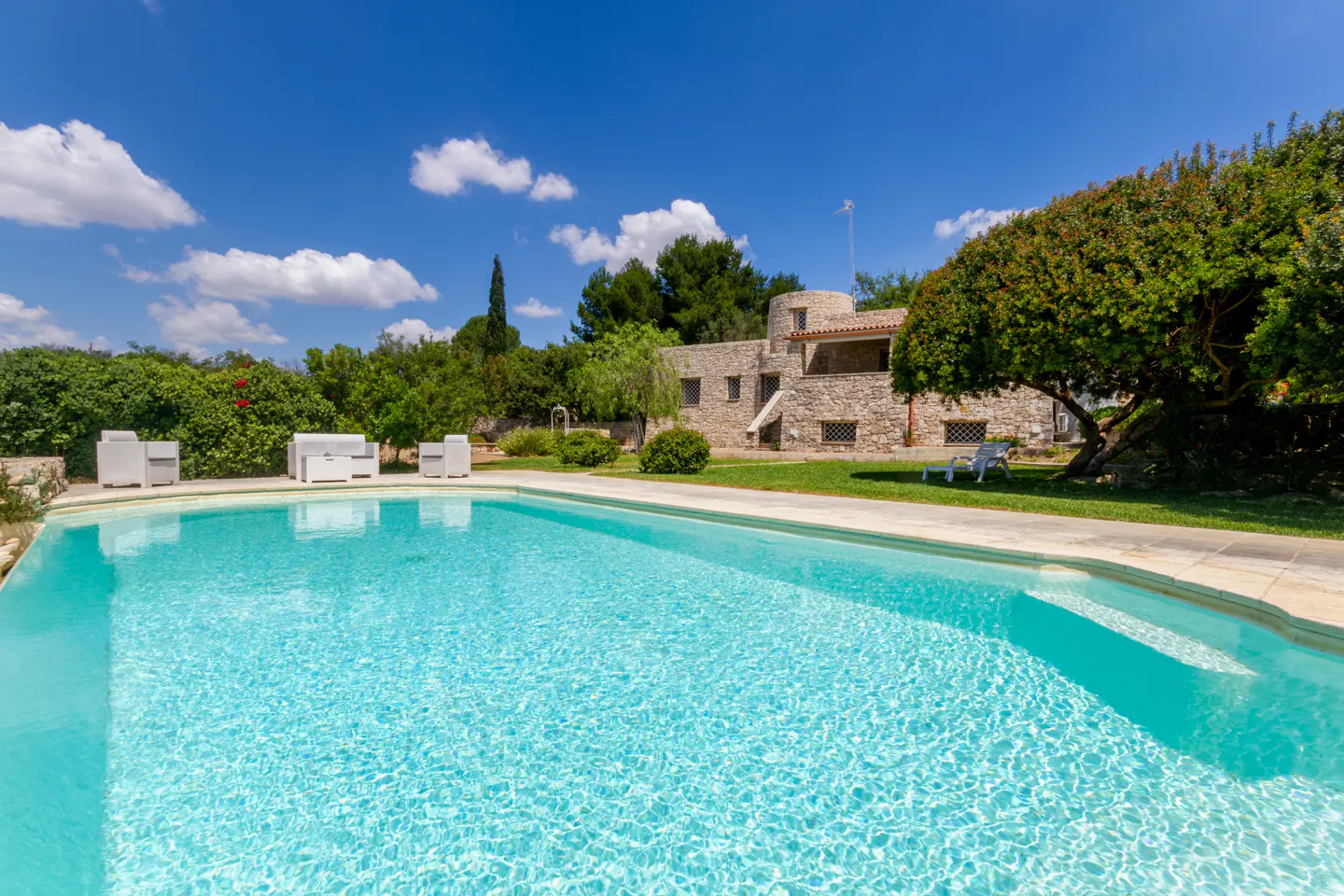 A turquoise swimming pool in front of a rustic stone villa with white patio furniture and lush green trees under a blue sky.