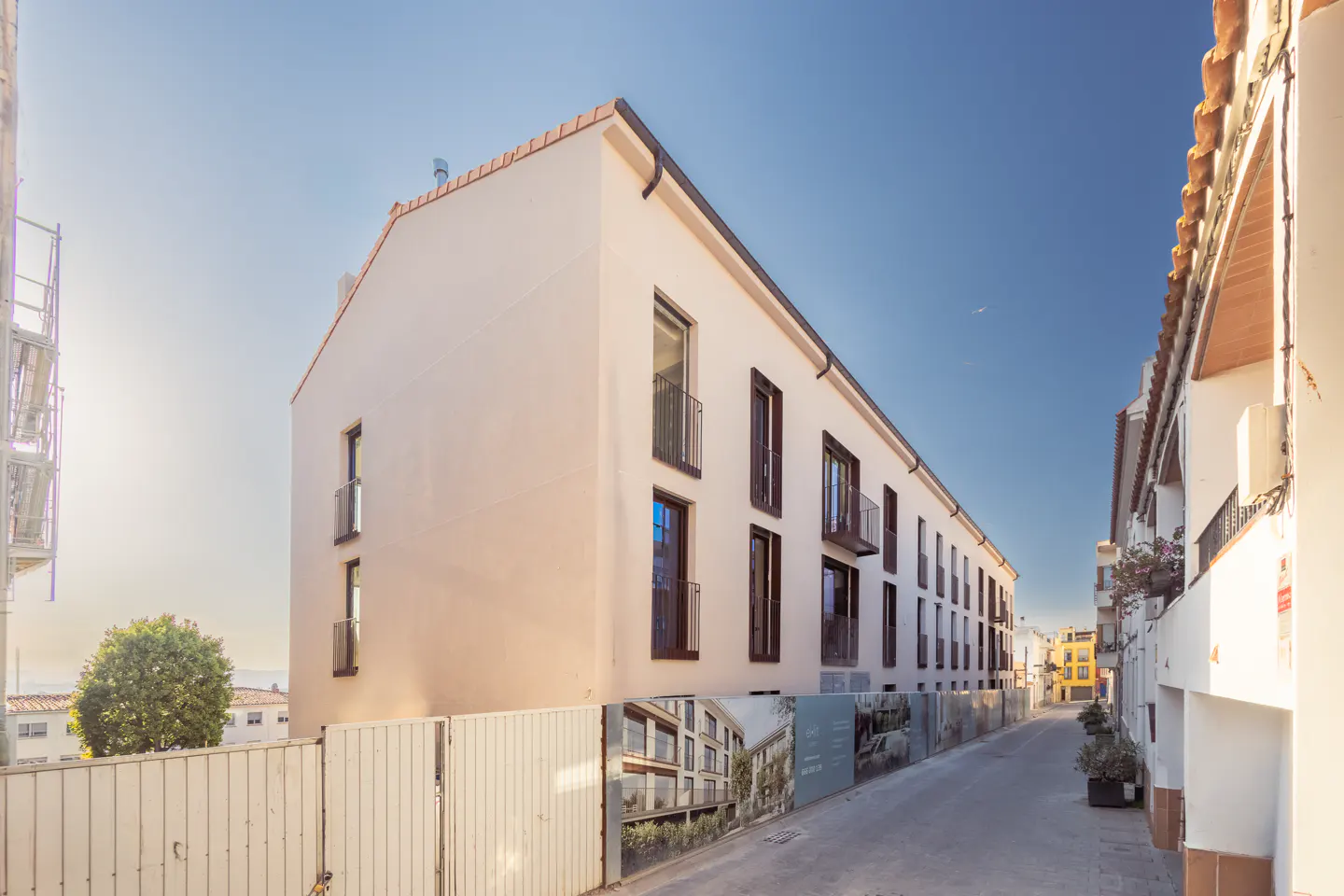 Exterior view of a long, narrow, white building with dark-framed windows and small balconies on a sunny street.