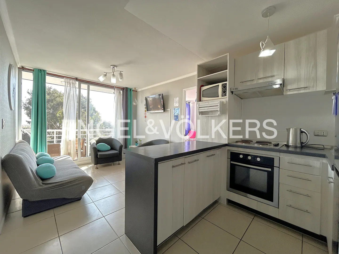Bright apartment interior with a gray sofa, blue pillows, and a kitchen island with white cabinets. Balcony with green curtains in the background.