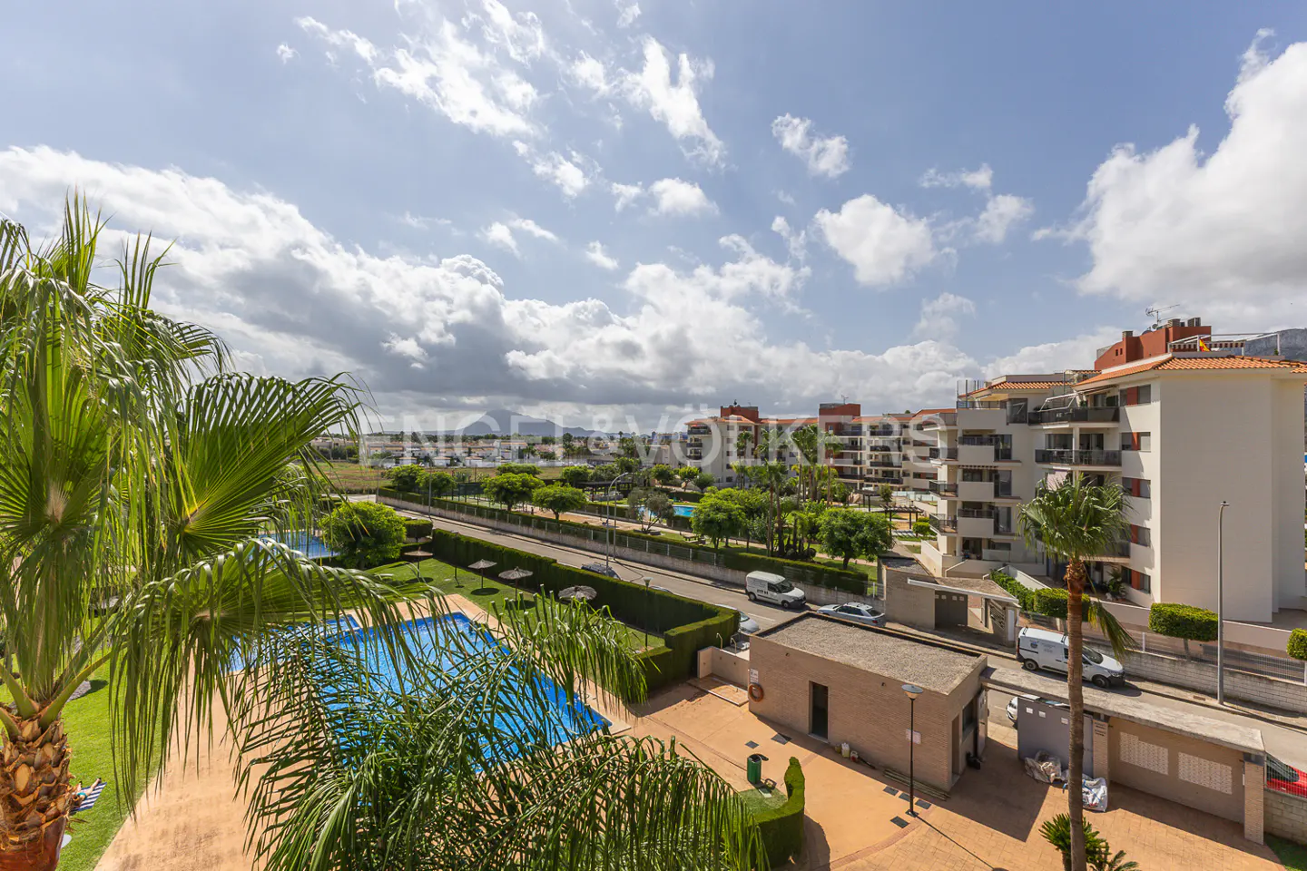 Aerial view of a blue swimming pool, palm trees, and residential buildings under a cloudy sky.