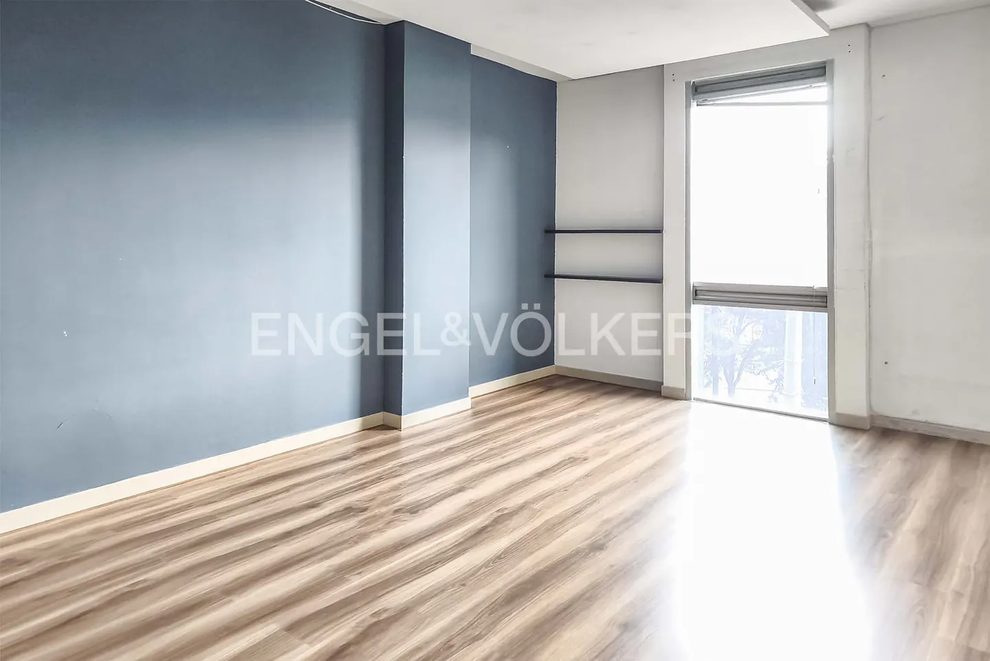 Empty room with blue walls, wood floor, and a large window with blinds. Engel & Völkers logo visible on the wall.