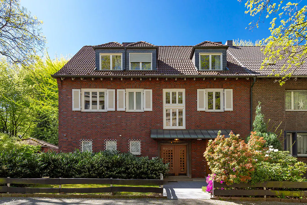Two-story brick house with a brown tiled roof, white window shutters, and a green hedge in front. Blue sky.