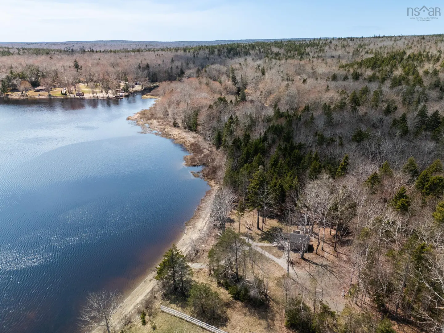 Aerial view of a lake bordered by trees, some bare and some evergreen, under a blue sky. A small building is visible in the trees.