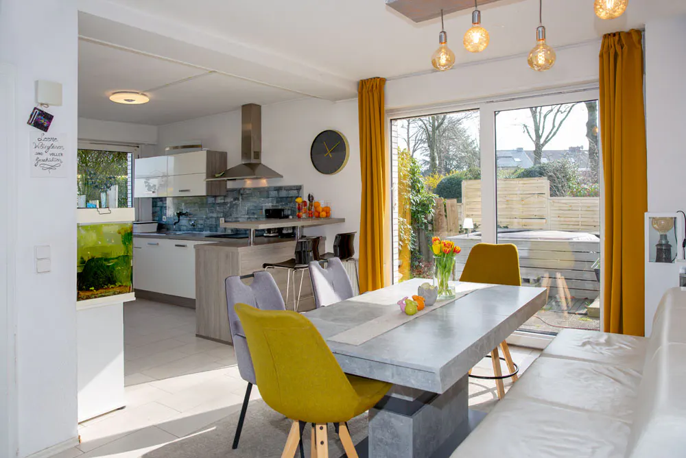 Bright, modern kitchen and dining area with yellow accents. A concrete table is surrounded by yellow and gray chairs. A kitchen and backyard are visible.