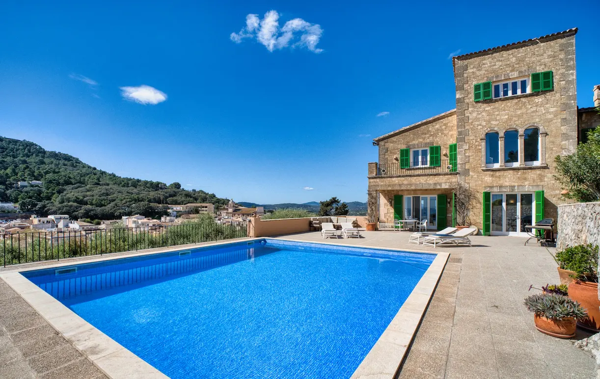 Luxury villa with a blue tiled pool, stone patio, and green shutters under a clear blue sky. Distant hills and town in the background.