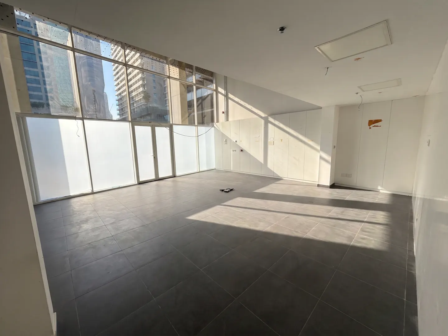 Empty office space with black tile floor, white walls, and large windows with frosted glass. Sunlight streams in, casting shadows across the room.