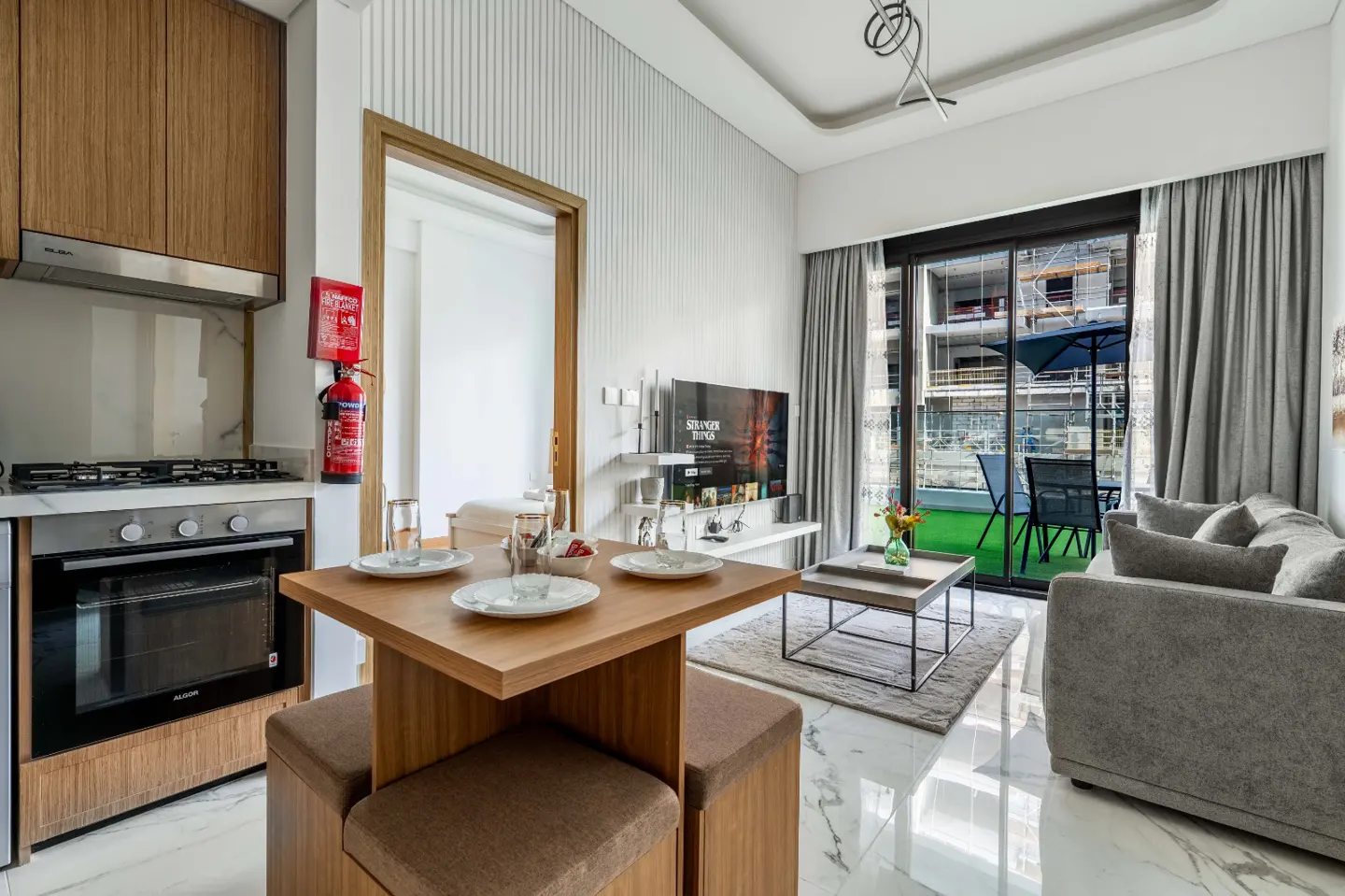 Modern studio apartment with a wooden dining set, grey sofa, kitchen area, and a balcony view through sliding glass doors.