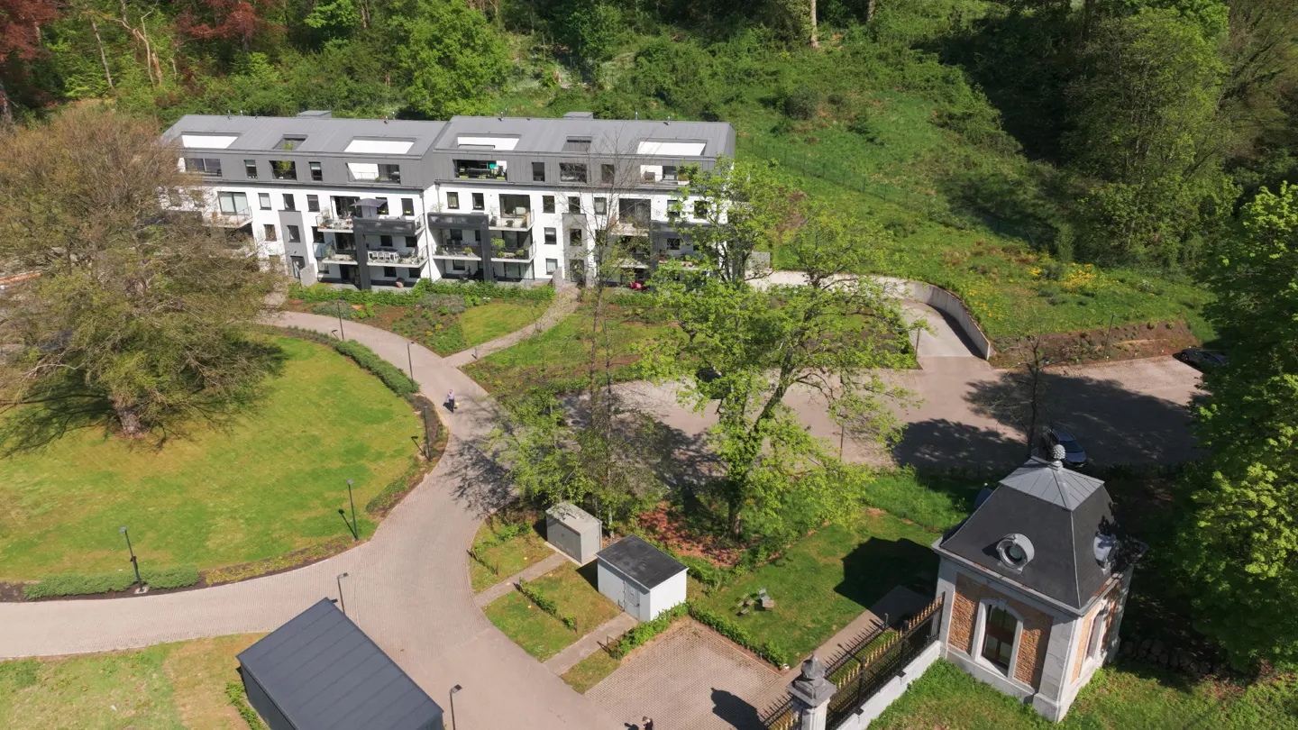 Aerial view of modern white apartments with gray roofs, surrounded by green lawns, trees, and a small brick building with a black roof.