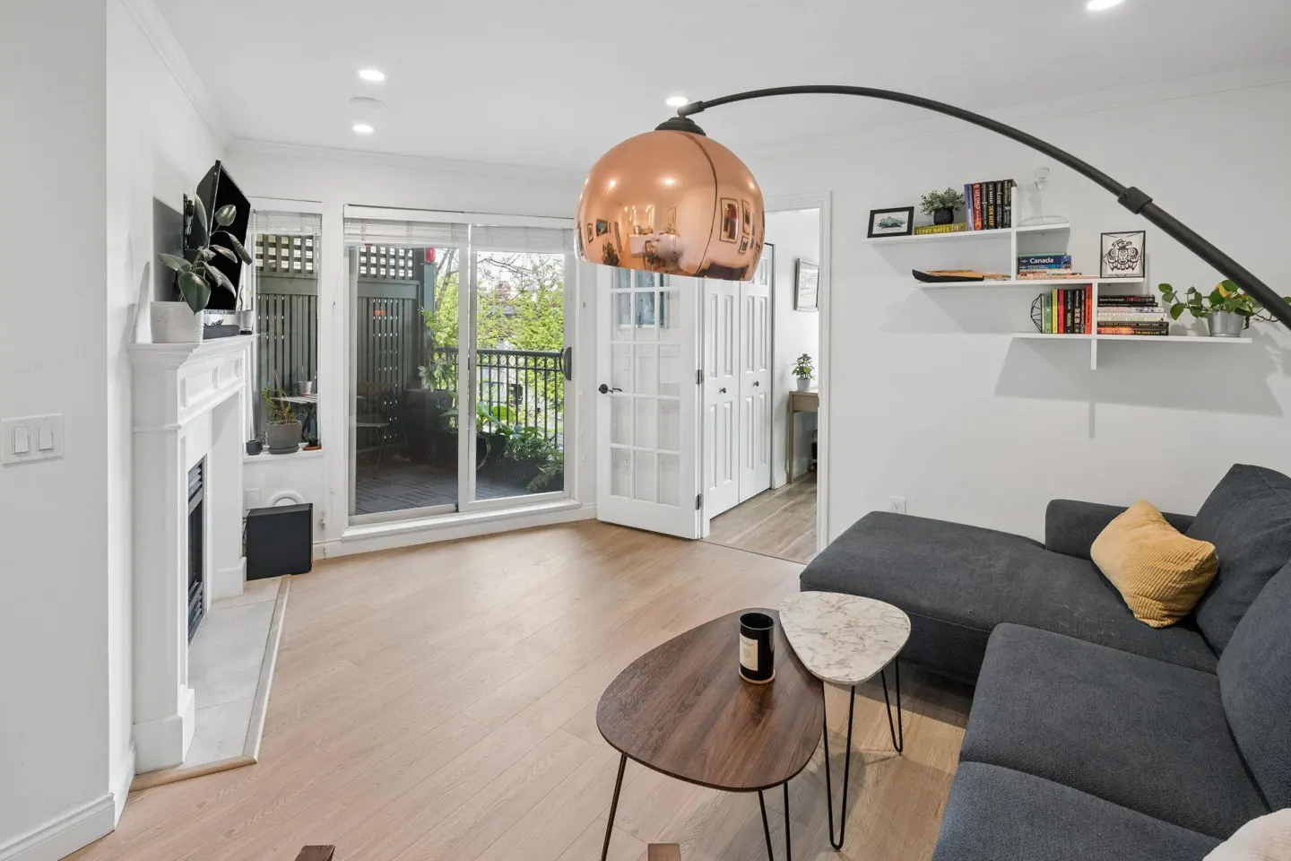 Bright living room with hardwood floors, a gray sectional sofa, and a white fireplace. Sliding glass doors lead to a balcony. A copper arc lamp hangs overhead.