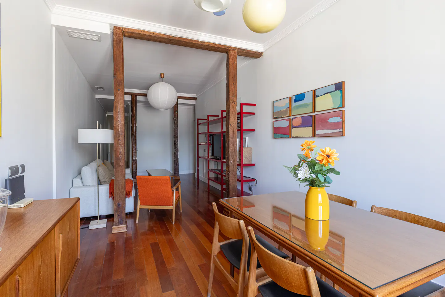 Modern dining area with glass table, yellow vase, and wooden chairs, leading to a living room with exposed wooden beams.