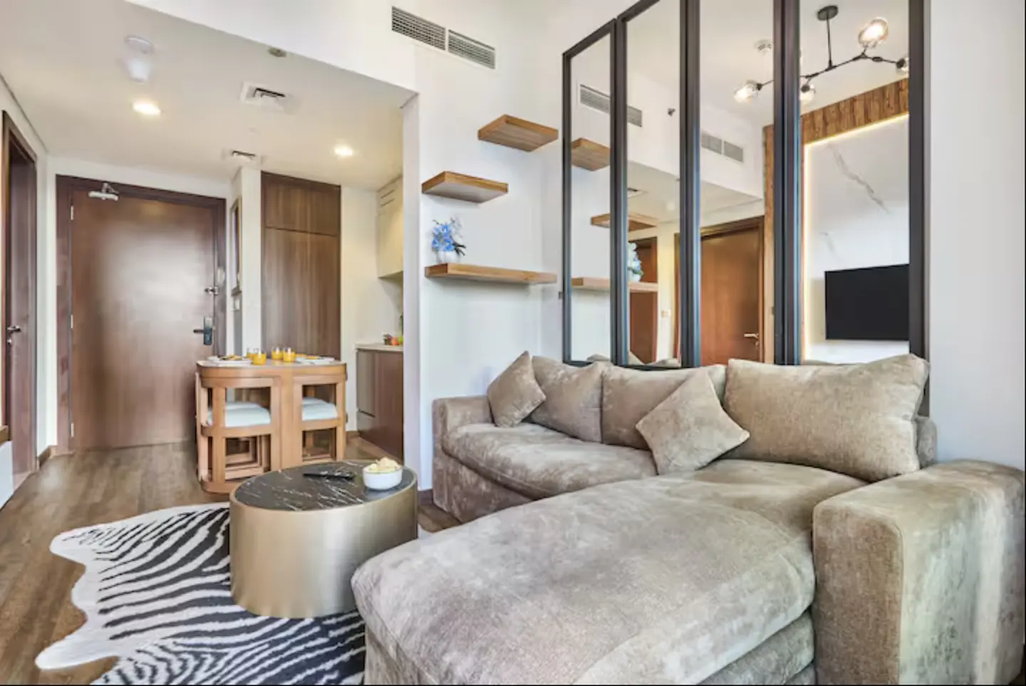 Interior view of a modern apartment with a brown sofa, zebra rug, and a round table. Shelves and mirrors add depth to the space.