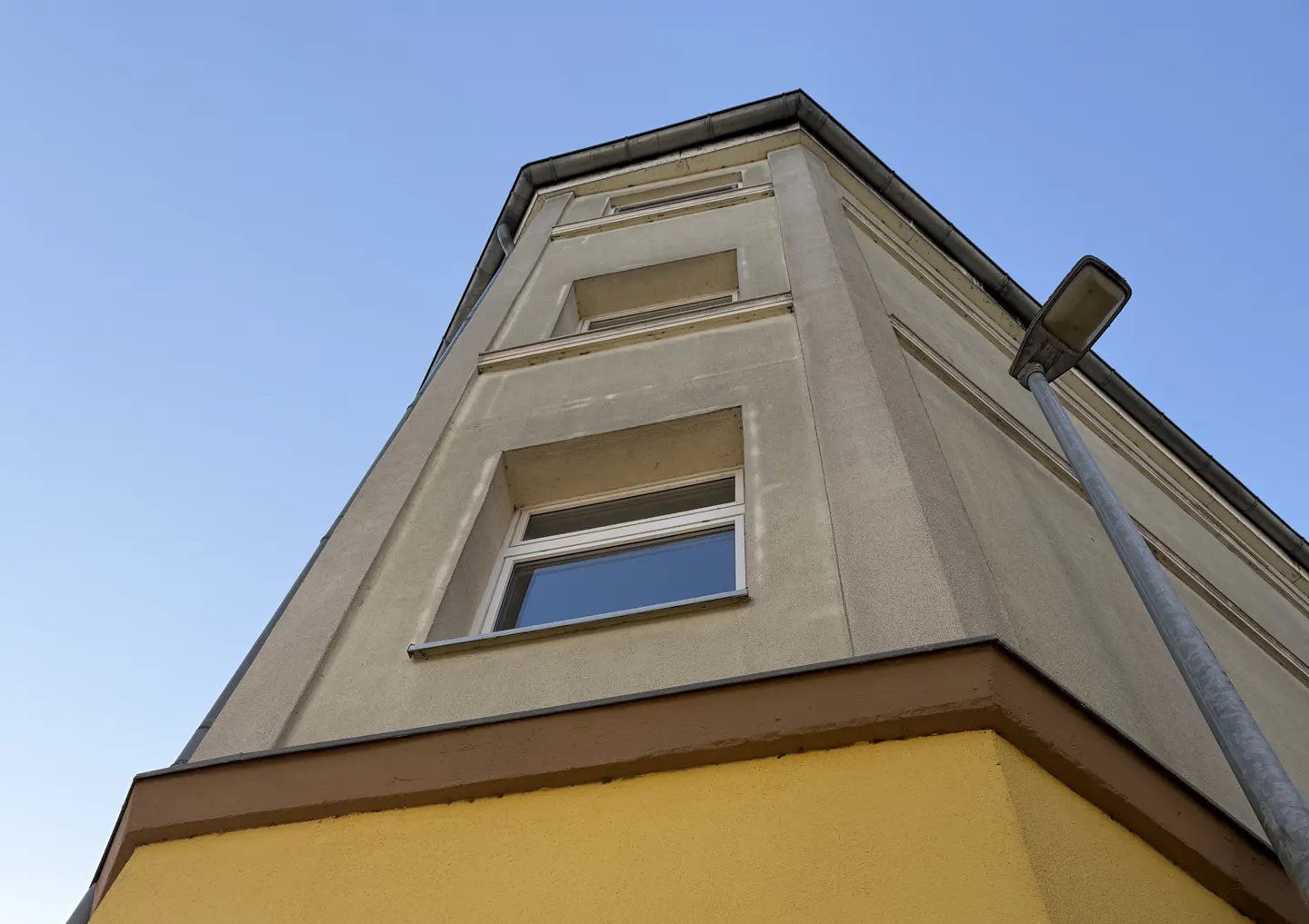 Low-angle shot of a building corner with yellow base, beige upper floors, white windows, and a street lamp against a blue sky.