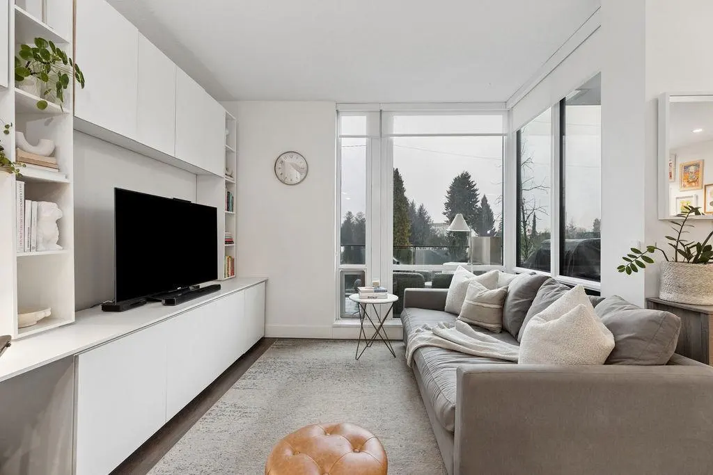 Bright living room with a gray sofa, white built-in shelves, and a large window overlooking trees. A brown ottoman sits on a patterned rug.