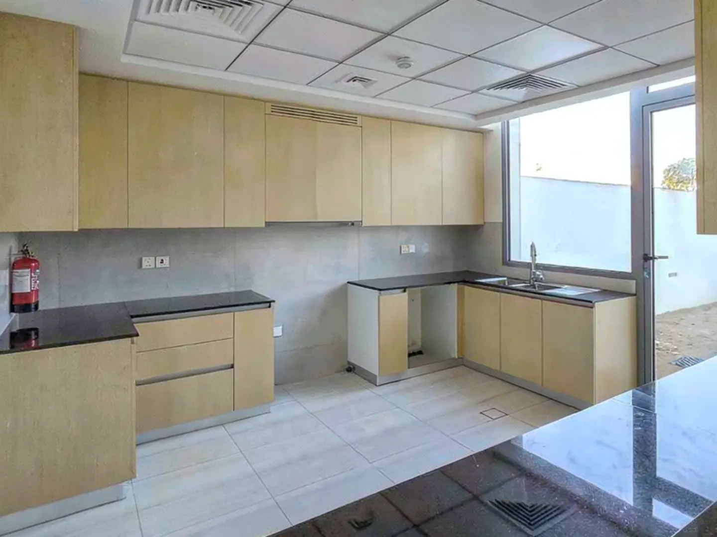 Bright kitchen with light wood cabinets, black countertops, and a large window. A red fire extinguisher is visible on the left wall.