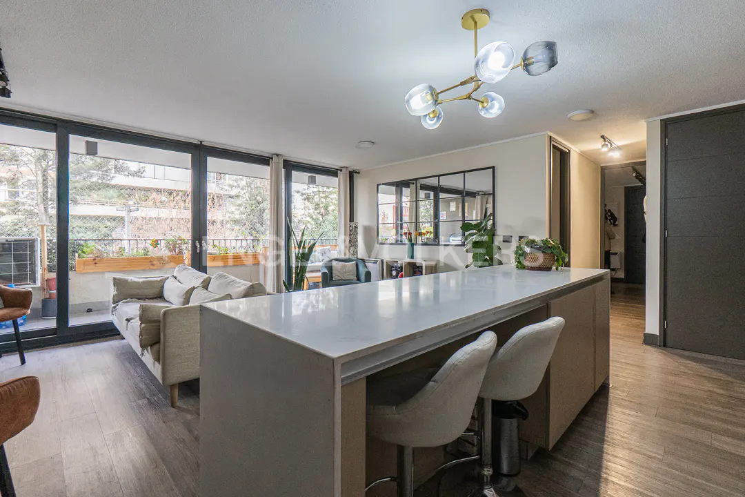 Modern open-plan living area with a white kitchen island, two bar stools, a beige sofa, and a large glass balcony door.