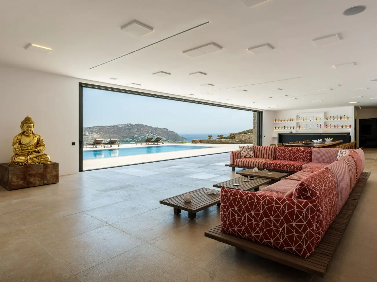 Bright living room with a red patterned sofa, wooden tables, and a golden Buddha statue. A pool and ocean view are visible through a large open window.