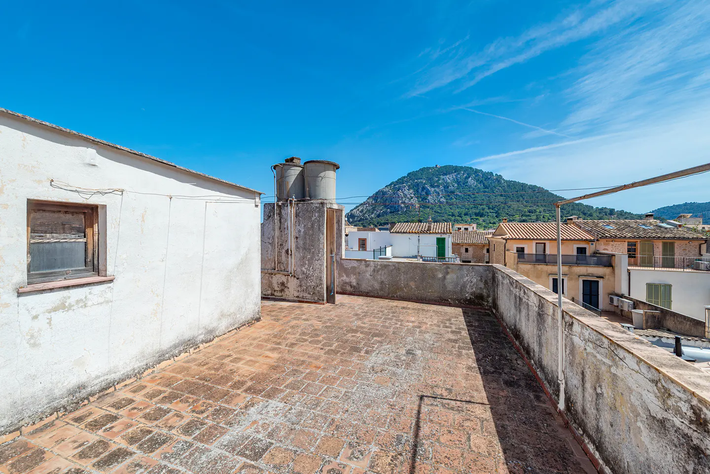 A rooftop view with brick tiles, a white building, water tanks, and a mountain backdrop under a blue sky.