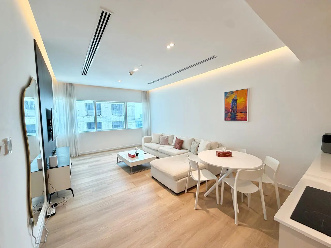 Bright, modern apartment interior with white walls, light wood floors, and a large white sectional sofa. A round white table with chairs sits nearby.