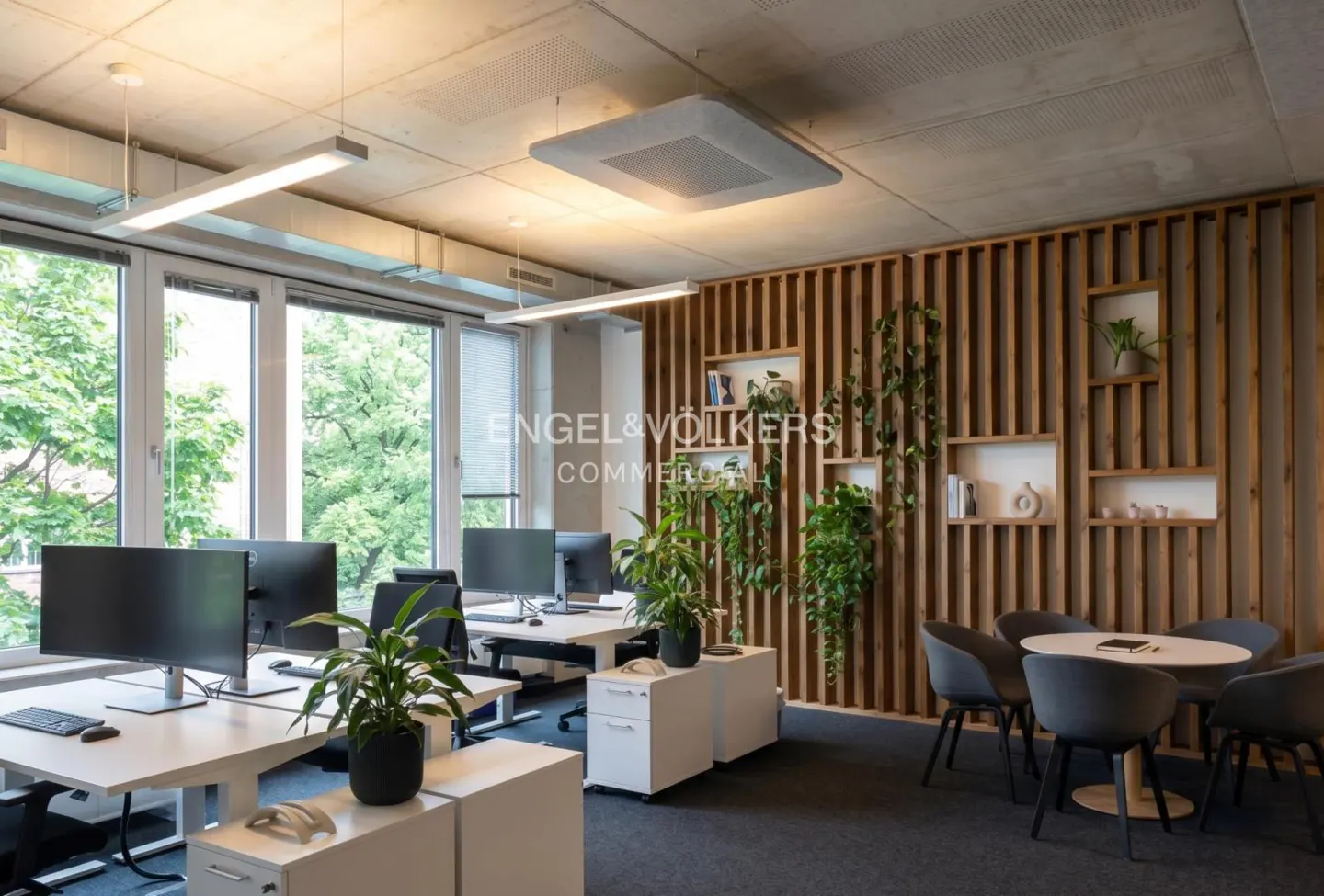 Bright office space with desks, computers, and plants. A wood slat wall with shelves and greenery adds a natural touch. A round table with chairs is in the corner.