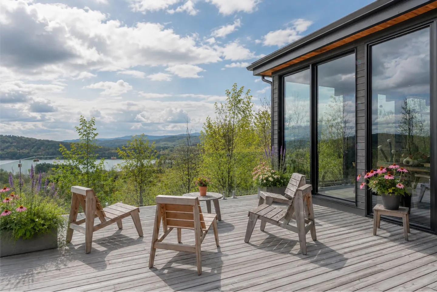 Modern wooden deck with lounge chairs and potted flowers overlooking a scenic lake and mountain landscape under a blue sky.