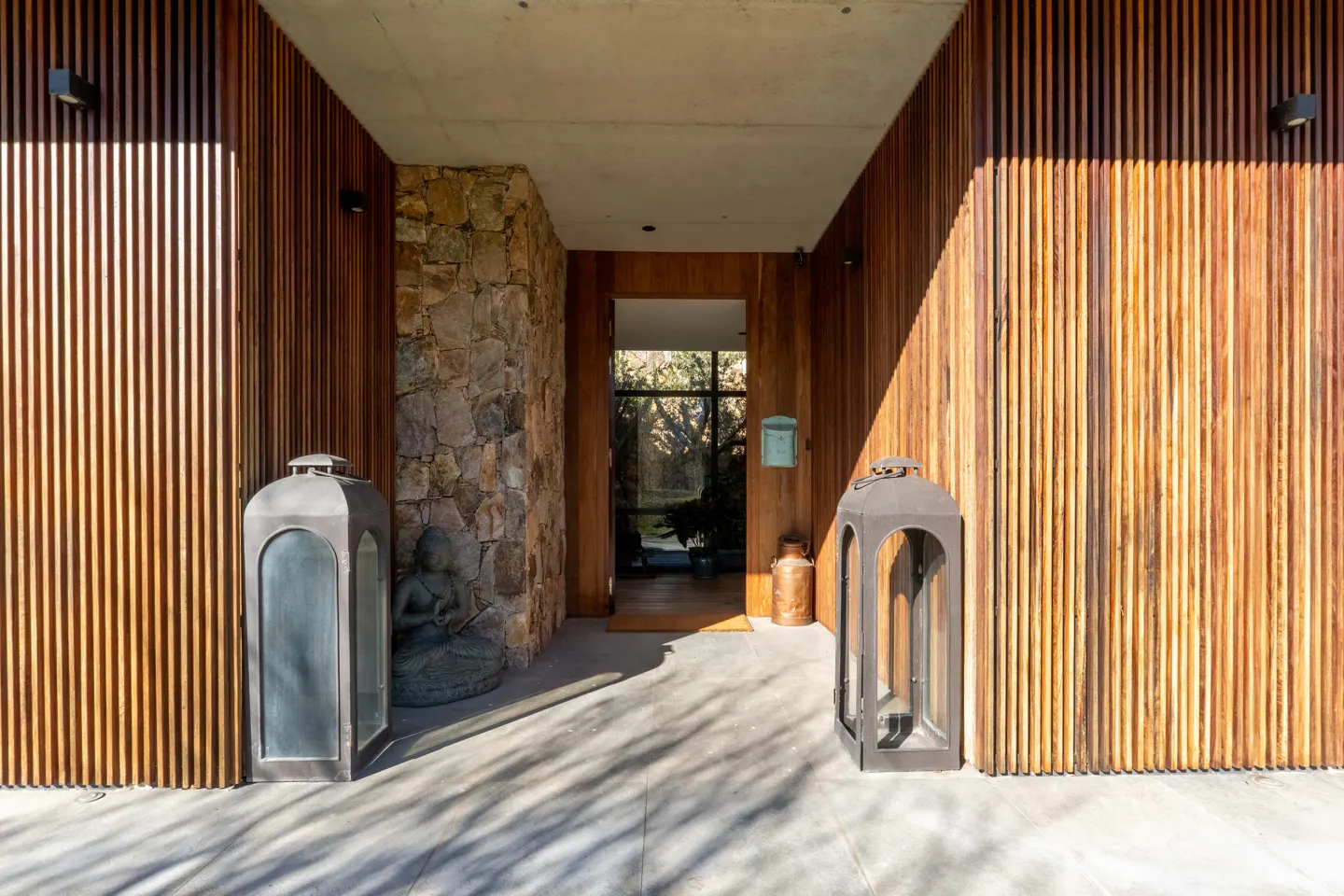 Modern entryway with vertical wood slats, stone accent wall, Buddha statue, and two large lanterns on a concrete porch.