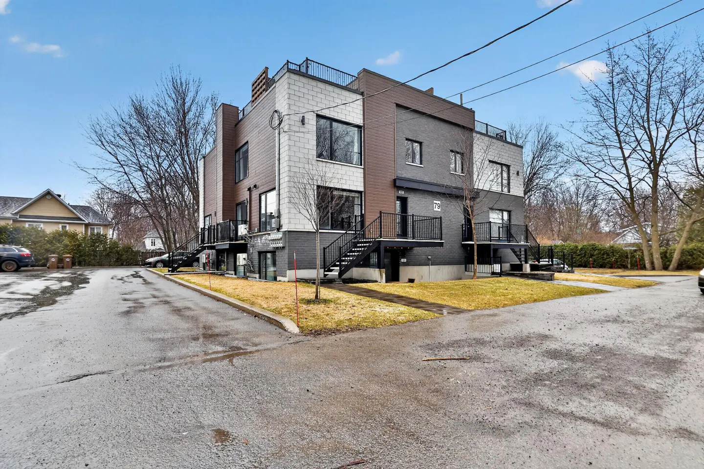 Modern three-story apartment building with gray brick, brown siding, and black balconies. A paved driveway leads to the building.